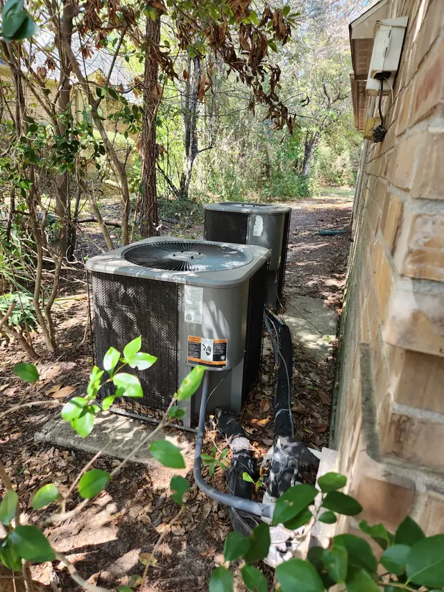 Two condensers on pads beside a brick home on a wooded lot